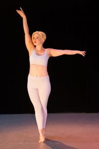 An image of Kelsey dancing. She stands on her tip-toes (sous-sus) with her arms up and out. 